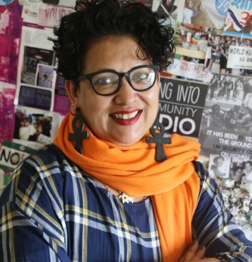 A woman with short curly dark hair smiling in front of a wall of photos, wearing a bright orange scarf