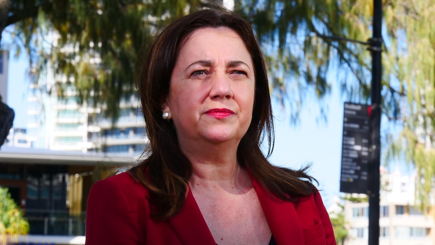 Premier in red jacket with dark brown hair looking out with trees, blue sky and buildings behind