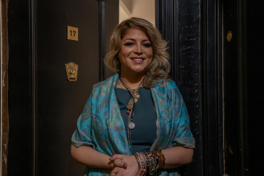 a woman in a blue silk scarfs smiles outside an apartment door
