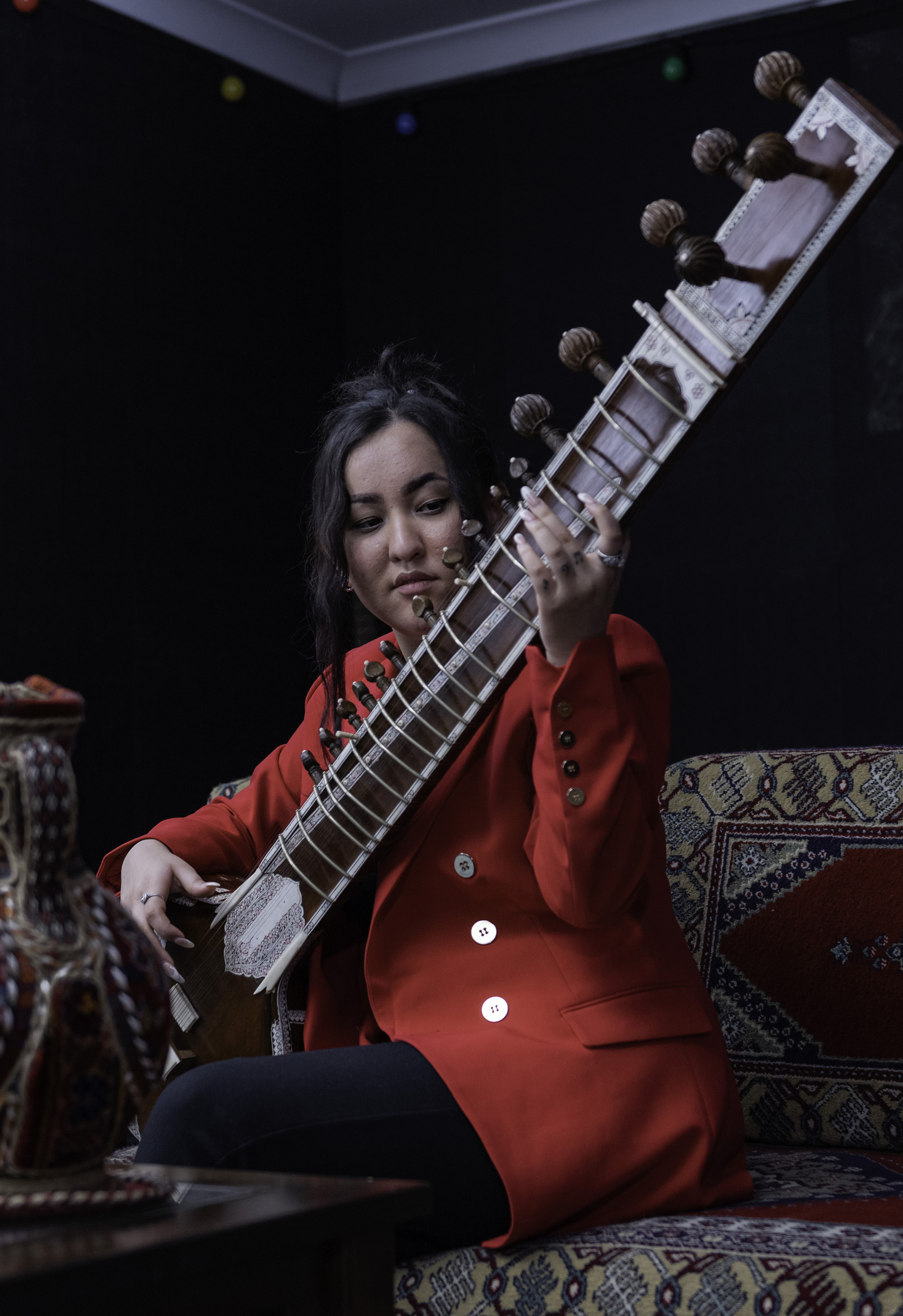 A young Afghan woman in a red jacket plays a sitar that is sitting in her lap