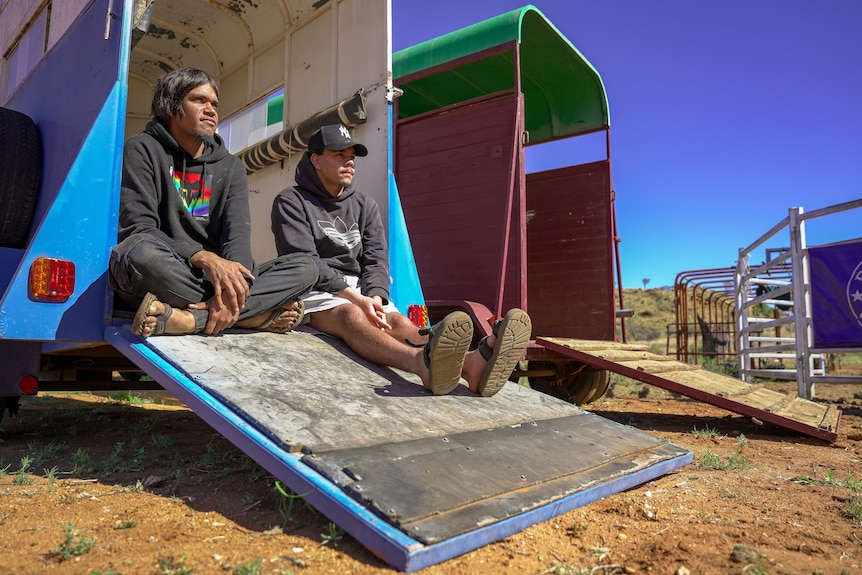 Two young men sit on the back of an open horse trailer, looking to the distance.