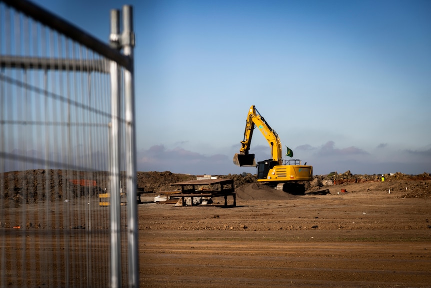 A yellow excavator is parked on a cleared area of dirt, next to metal construction site fencing.