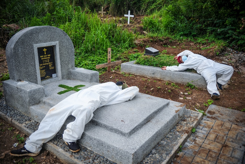 Two men in full PPE lie across graves in a cemetery 
