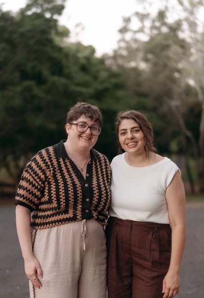 Two sisters smiling at the camera in front of a small forest.