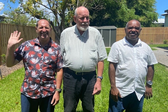 three men standing in the sun in a backyard, one is waving