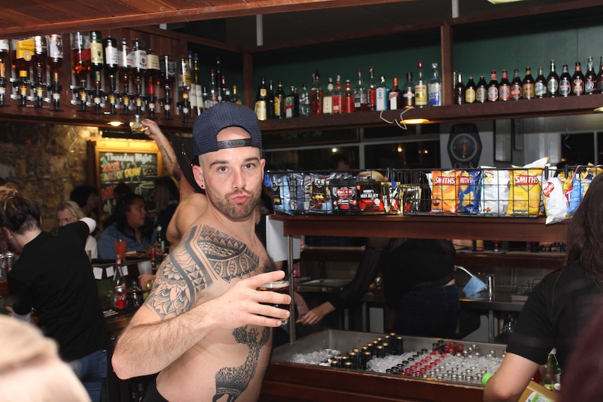 A tattooed male bartender wearing no shirt strikes a pose behind the bar of am pub.