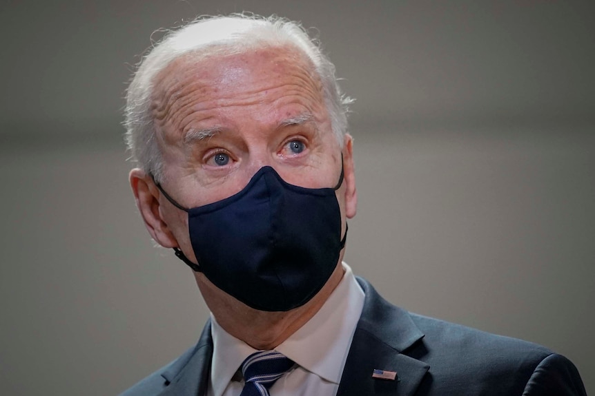 An elderly man with white hair wearing a suit and a mask.