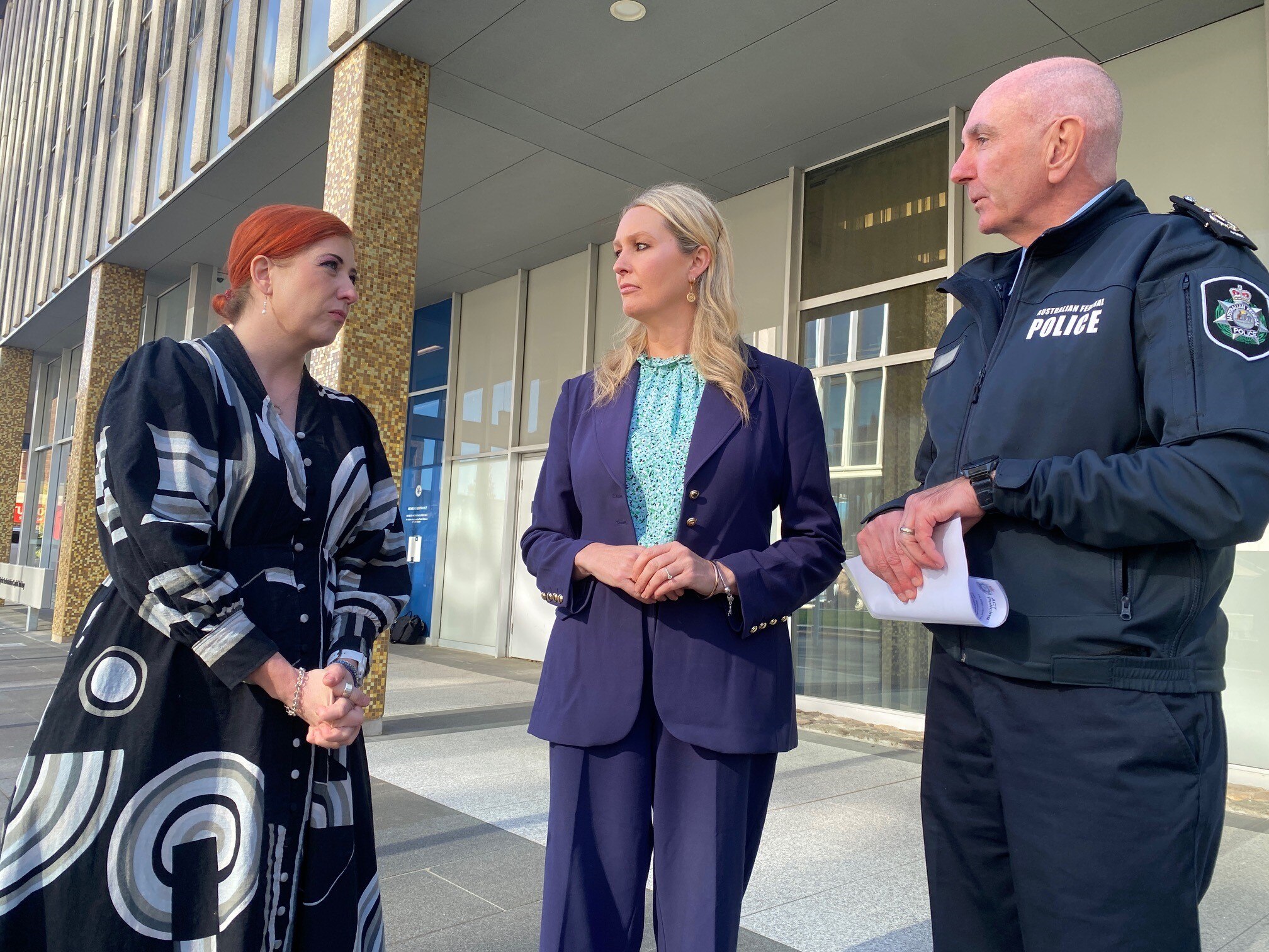Three people stand in a semi circle, a woman with orange hair, a woman in a indigo suit and a bald man. 