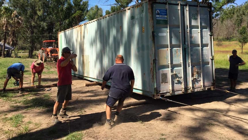 a shipping container with logs under it is moved on ropes with men around it chceking it progress