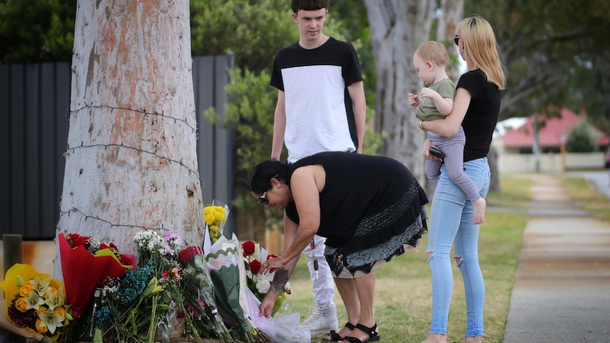 A woman bends down to lay flowers by a tree, as a young man, woman and baby watch on