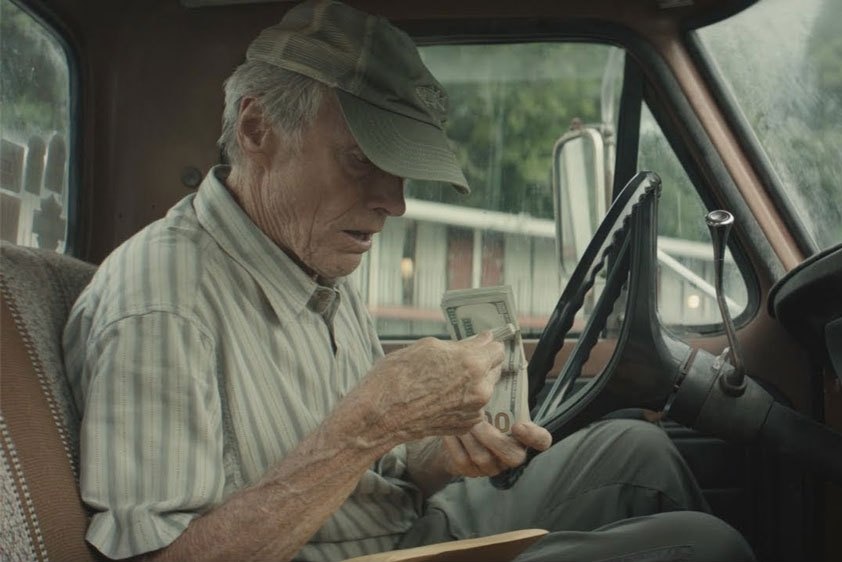 Clint Eastwood counting money and wearing a cap in a pick-up truck, in the film The Mule.