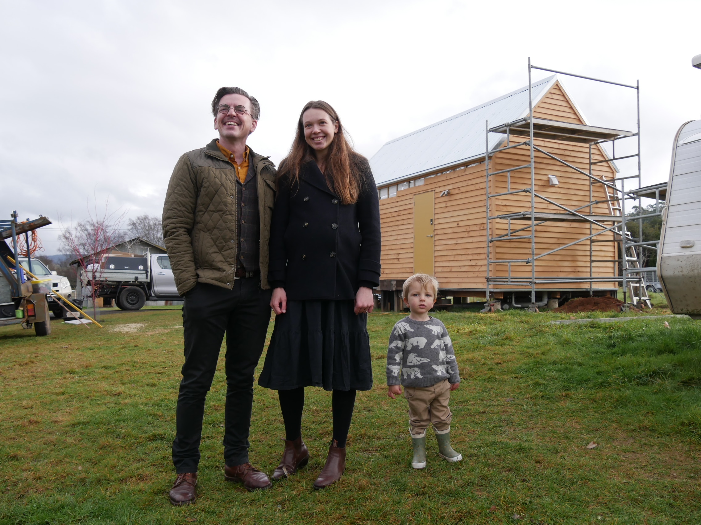 A family of three standing in front of a small home they are building.