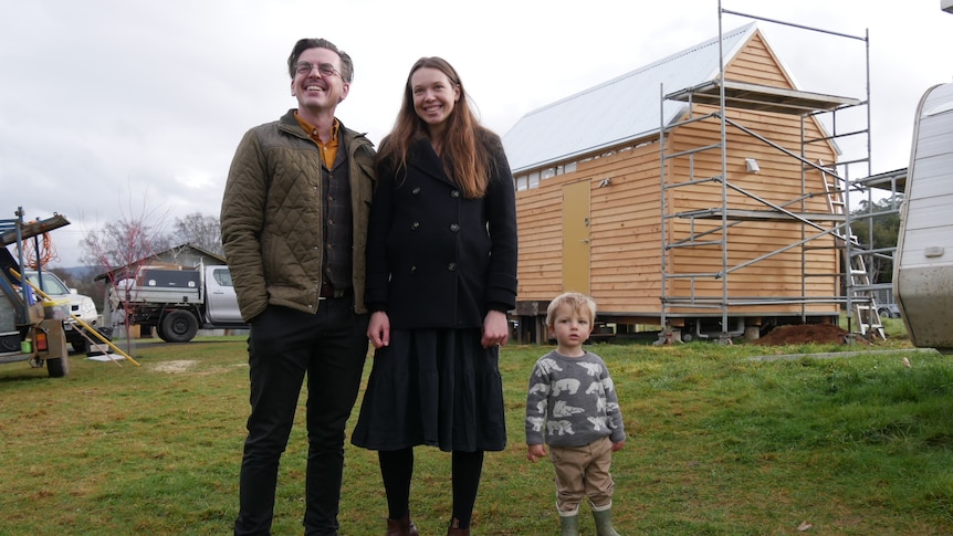 A family of three standing in front of a small home they are building.