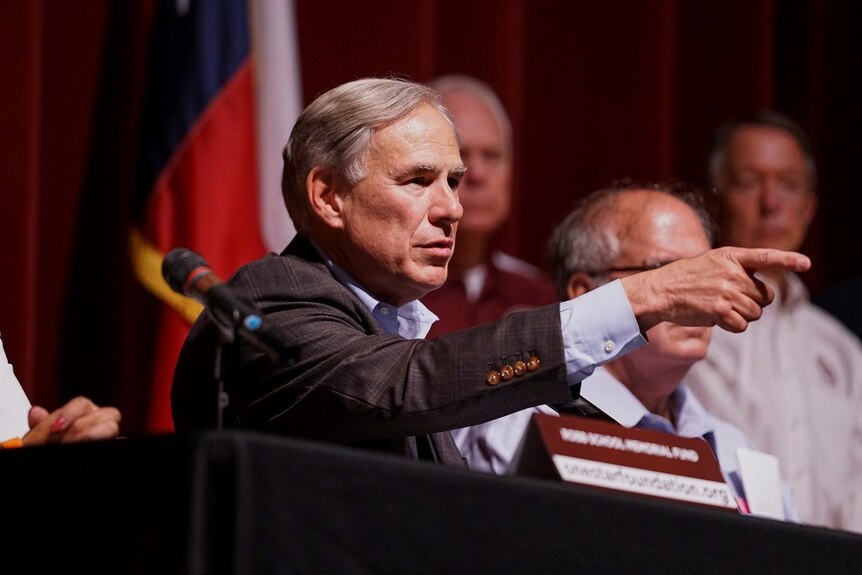 A middle-aged white man in a checked suit points while seated behind a table at a news conference.