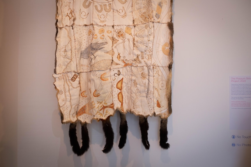 A possum skin cloak etched with artwork of animals and places, with furry dark tails dangling from the bottom.