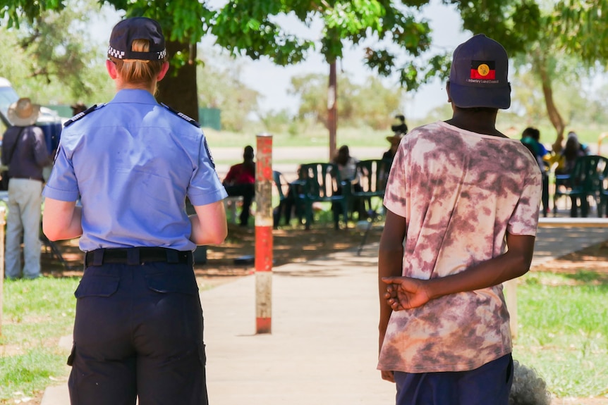 A police officer and boy, both viewed from behind, at a community meeting. Officer in uniform, boy in brightly patterned shirt