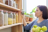 Woman with bowl of green apples puts a jar on a pantry shelf.