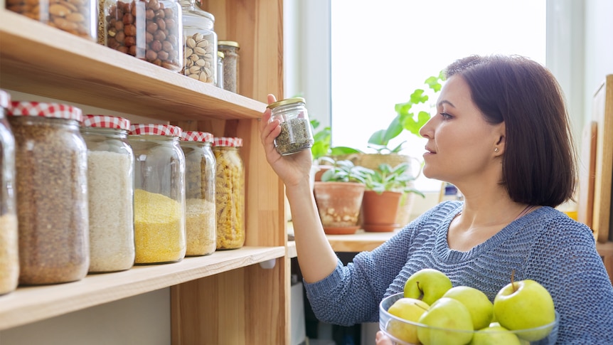 Woman with bowl of green apples puts a jar on a pantry shelf.