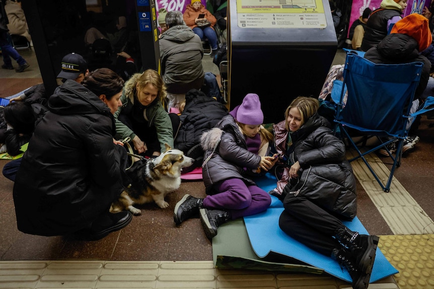 A family bundled up in coats sits on the floor of a train station with their pet corgi