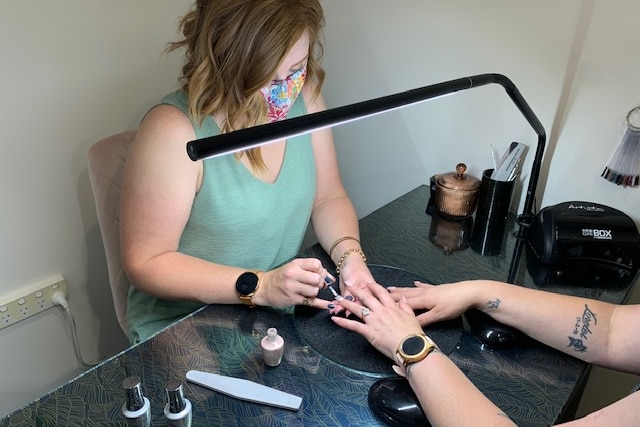 A woman in a mask paints the nails of another woman under a light