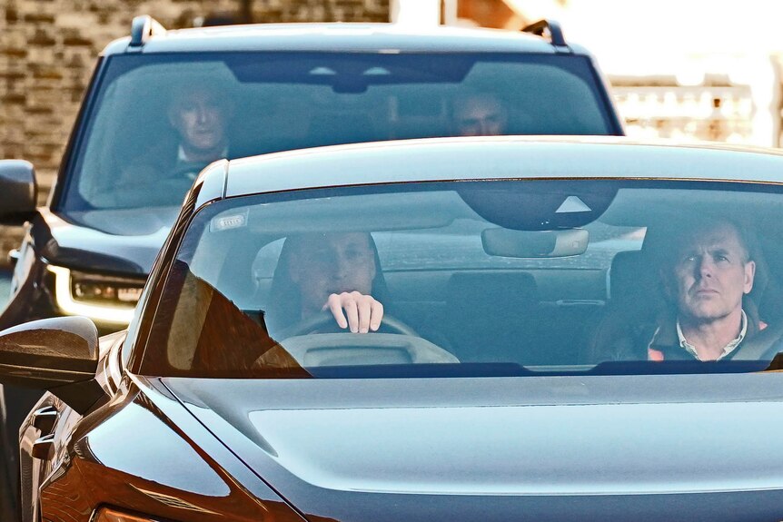 A zoomed-in photo of Prince William driving a car with a man in the passenger seat and another car following