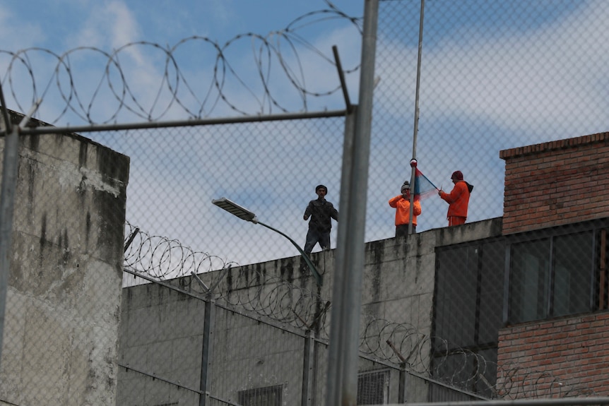 Three men in street clothes stand on top of the roof of a concrete building, photographed through barbed wire.