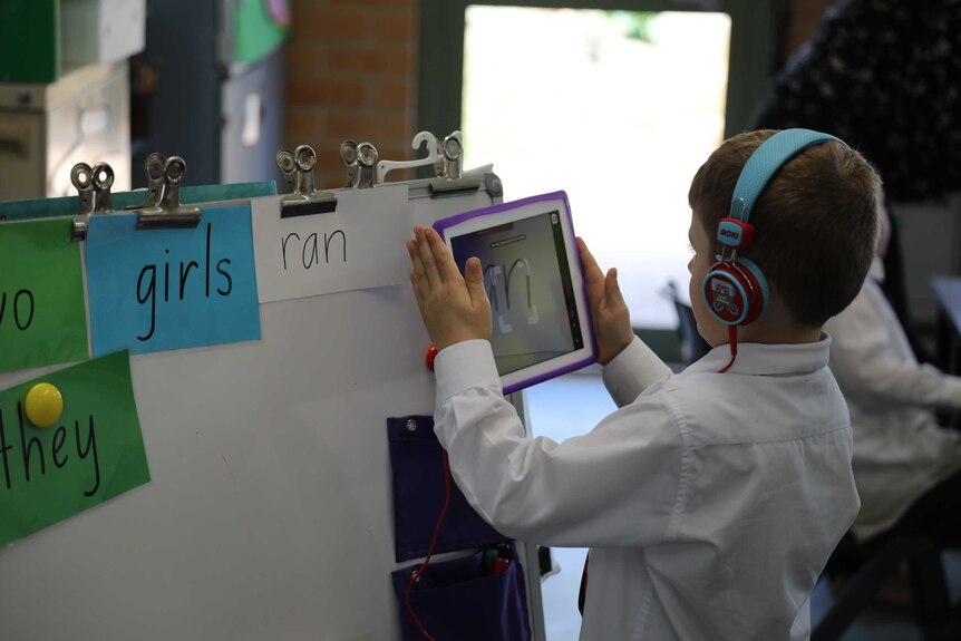 A primary school student stands holding an ipad up near words on a whiteboard.