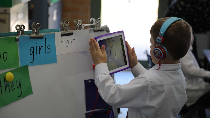 A primary school student stands holding an ipad up near words on a whiteboard.
