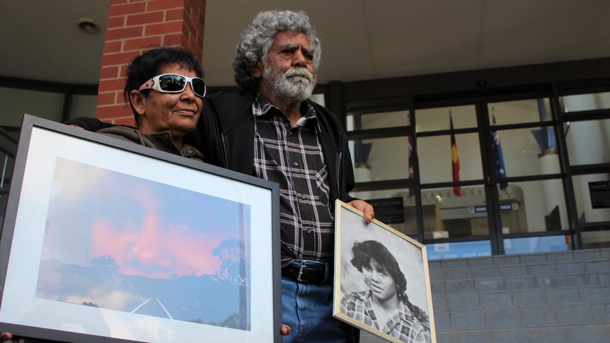Two aboriginal elders stand on the front of court house steps holding each other and the photo of their dead family member