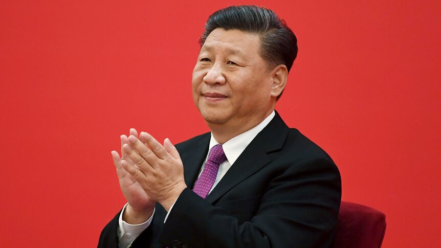 A man in a suit smiles and claps as he sits before a red background.