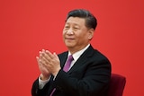 A man in a suit smiles and claps as he sits before a red background.