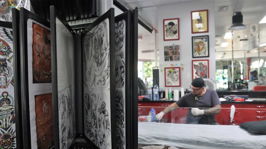 A rack of posters displaying tattoo patterns with a tattoo artist filling up his inks in the background.