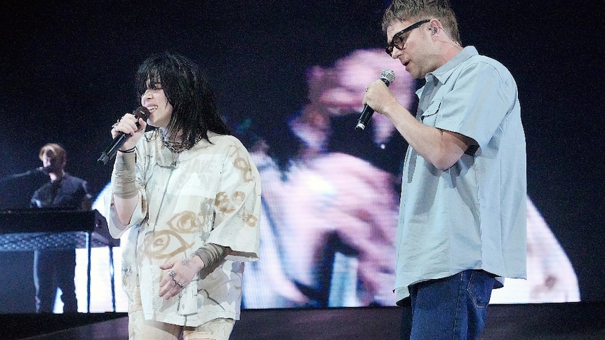 Billie Eilish, dressed in beige with black hair, performing with Damon Albarn, dressed in blue shirt and jeans