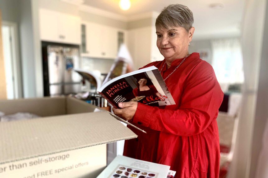 A woman looks at a cook book, with a pile of books and a cardboard box in front of her.