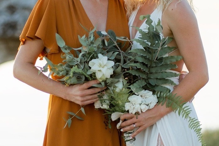 A bridesmaid in a copper dress, next to a bride in white, clutching a bouquet of white lily-like turmeric flowers.