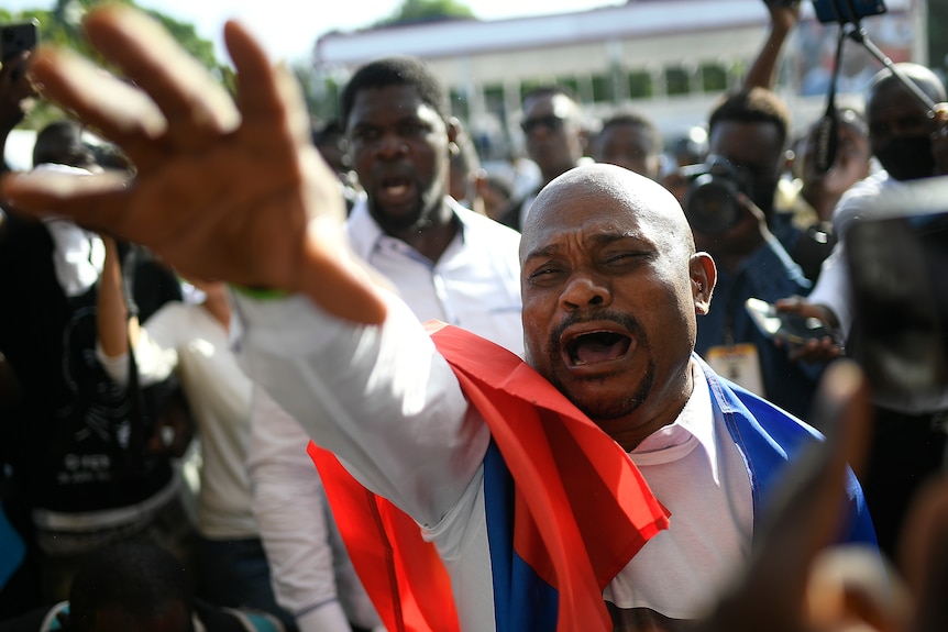 a man crying with hands up and haiti flag on his back in crowd of protestors