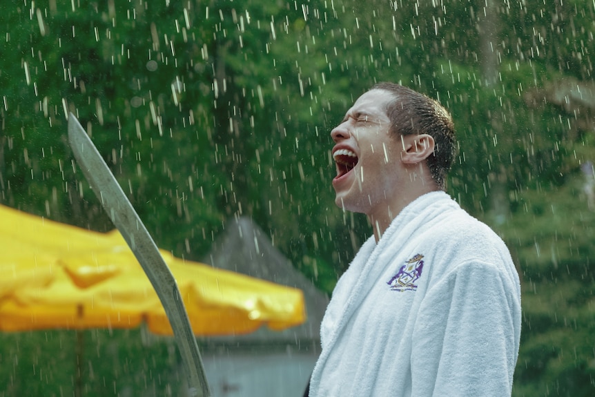 Bărbat alb purtând un halat de baie alb ținând o sabie țipând pasional în ploaie.  