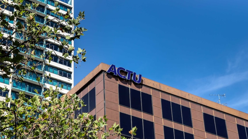 ACTU sign at top corner of brown office building, with tree branches in foreground and large office building in background.