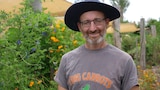 Ryko smiling at the camera wearing a dark wide brimmed hat, "i dig carrots" t-shirt with flowers to his left.