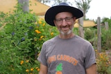 Ryko smiling at the camera wearing a dark wide brimmed hat, "i dig carrots" t-shirt with flowers to his left.