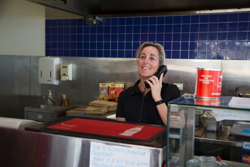 A blond woman answers the phone from behind a takeaway counter. A blue tiled industrial kitchen is behind her.