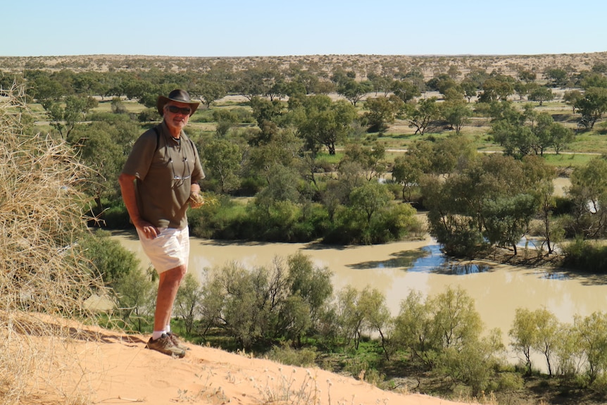 A man in a green shirt and white shorts stands on red dirt beside a brown river lined with green trees.