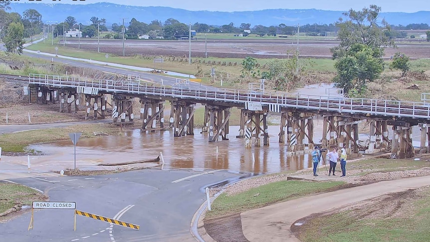 Camera crew interviewing a person beside flooded road under a bridge.