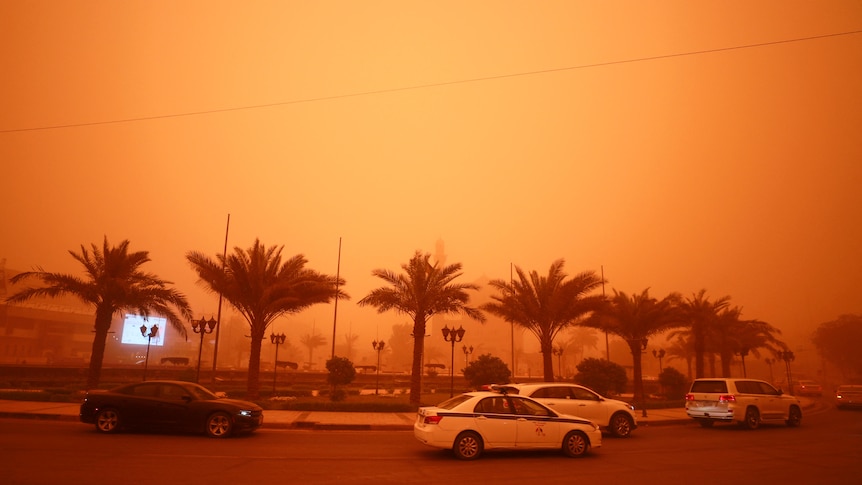 Vehicles drive along a road during a severe dust storm under an orange sky.