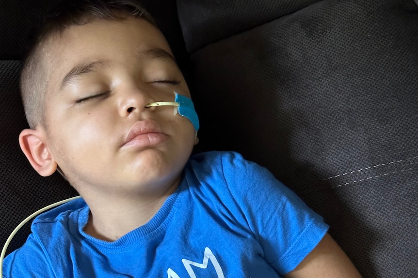 A young boy in a blue t-shirt sleeps in a hospital bed. He has a tube extending from his nose