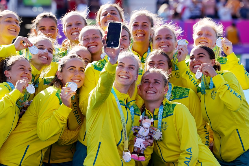 A group of Australian women's hockey players smile up at the camera as they take a selfie with their silver medals.