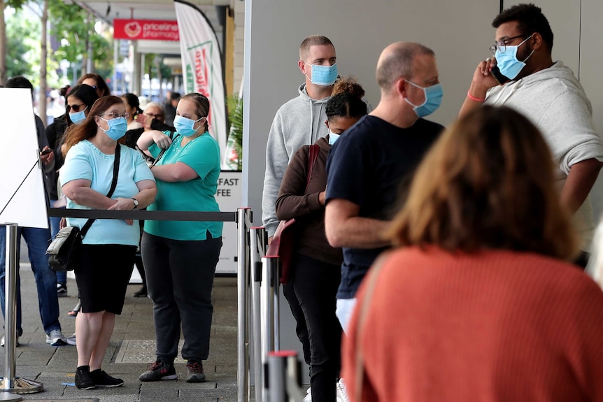 A group of people, some wearing face masks, stand in line outside a retail store. they are socially distanced by barriers