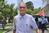 A slim elderly man in light blue shirt and glasses walks towards the camera outside a building on a sunny day