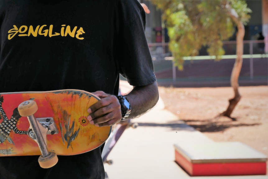 A close-up of a person wearing a black tshirt that says songlines holding a skateboard painted red and orange.