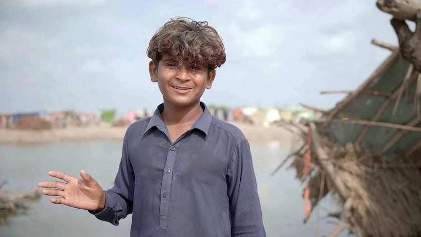 A boy smiles and waves to camera. Across a body of water there is makeshift tent dwellings.